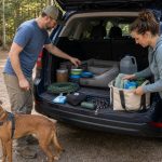 Dog owners organizing dog camping gear at an SUV tailgate with a harnessed dog at a wooded campground