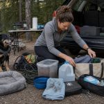 Dog owner reorganizing messy dog camping gear beside an SUV at a wooded campsite while a harnessed dog waits nearby